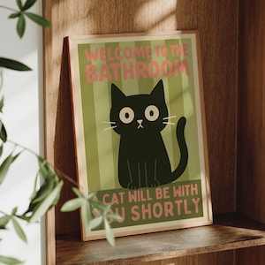 Può includere: Una stampa incorniciata con un'illustrazione di un gatto nero e il testo "WELCOME TO THE BATHROOM" e "CAT WILL BE WITH YOU SHORTLY". La stampa è in una cornice di legno ed è esposta su una mensola di legno.