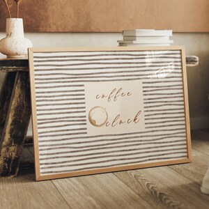 May include: A framed print with a light wood frame. The print has a striped background with the words "coffee o'clock" and a coffee ring stain. A vase with dried flowers sits on a wooden bench.