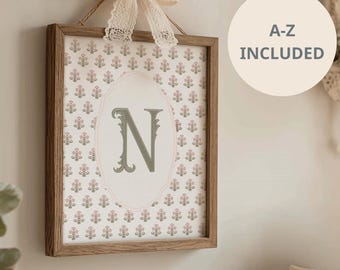 May include: A square wooden frame containing a print with a repeating floral pattern in muted pink and green tones. A central oval frame highlights the letter N in a dark green serif font. A cream lace bow is attached to the top. Text reads A-Z INCLUDED.
