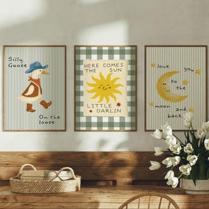 Pode incluir: Três impressões de arte emolduradas com ilustrações caprichosas. Uma apresenta um ganso com chapéu, outra um sol com texto e a última uma lua com estrelas. As impressões têm um fundo listrado ou xadrez e são exibidas em uma cômoda de madeira.