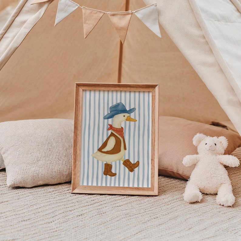 May include: A framed print featuring a cartoon duck dressed as a cowboy, wearing a blue hat, red bandana, brown vest, and boots. The background has blue and white vertical stripes. The frame is made of wood.