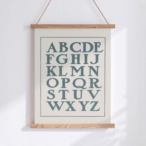 May include: A framed alphabet chart with the letters A through Z in a teal color. The poster is suspended by a natural rope and held in place by wooden bars at the top and bottom. The background is a light cream color.