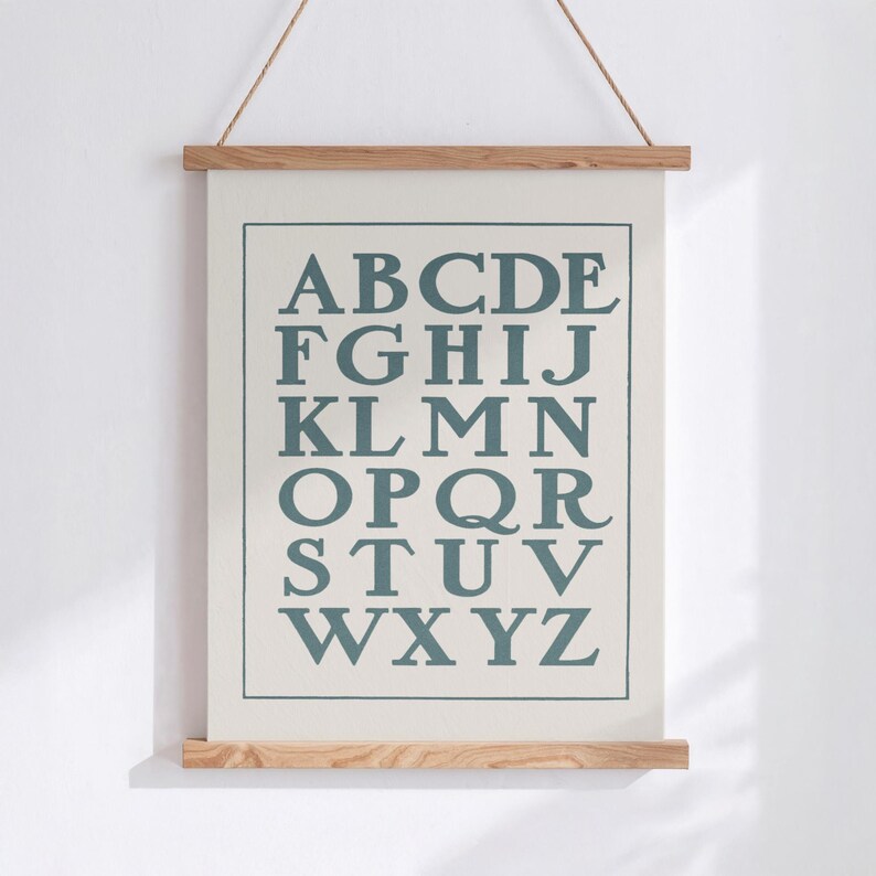 May include: A framed alphabet chart with the letters A through Z in a teal color. The poster is suspended by a natural rope and held in place by wooden bars at the top and bottom. The background is a light cream color.