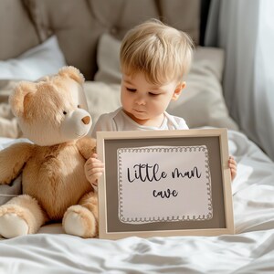 May include: A young child holding a framed sign that reads "Little man cave." A plush teddy bear sits to the child's left. The sign has a light brown frame and a white background with decorative border.