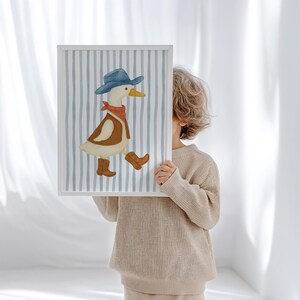 May include: A framed art print featuring a whimsical illustration of a duck dressed as a cowboy. The duck wears a blue hat, red bandana, brown vest, and cowboy boots, set against a blue and white striped background. The artwork is held by a person.