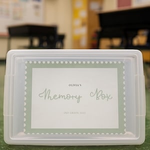 Può includere: Una scatola di memoria in plastica trasparente con un'etichetta bianca con la scritta "Olivia's Memory Box 2nd Grade 2023" in un carattere decorativo. La scatola è rettangolare con un coperchio incernierato.