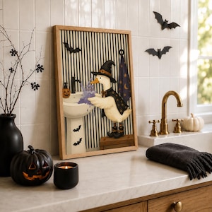 May include: A framed wall art print featuring a white duck wearing a black witch hat and cape, standing on a wooden crate to wash its wings in a pedestal sink. The background has black and white vertical stripes with bat silhouettes.