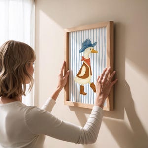 May include: A framed artwork featuring a whimsical illustration of a duck dressed as a cowboy. The duck wears a blue hat, red bandana, brown vest, and boots. The background has blue and white vertical stripes. The frame is made of light-colored wood.