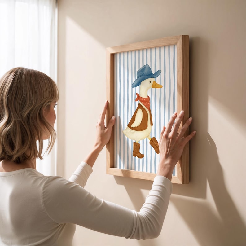 May include: A framed artwork featuring a whimsical illustration of a duck dressed as a cowboy. The duck wears a blue hat, red bandana, brown vest, and boots. The background has blue and white vertical stripes. The frame is made of light-colored wood.