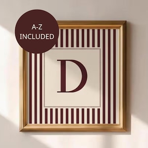 Puede incluir: Una impresión enmarcada con la letra "D" en borgoña sobre un fondo crema. La impresión está rodeada por un marco dorado y un patrón de rayas borgoña y crema. El texto "A-Z INCLUDED" está en un círculo marrón oscuro.