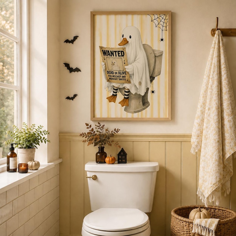 May include: A framed wall art print featuring a white ghost duck sitting on a toilet, holding a WANTED poster that reads DEAD OR ALIVE FOR MISCHIEF AND MIDNIGHT SNACKS. The background has yellow vertical stripes, a spider web, and small spiders.