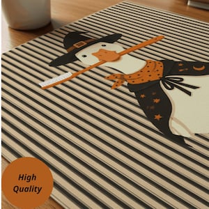 May include: A rectangular mat featuring a striped pattern in black and beige. The central graphic depicts a white goose wearing a black witch hat, an orange polka-dot bandana, and a black cape adorned with orange stars and a crescent moon.