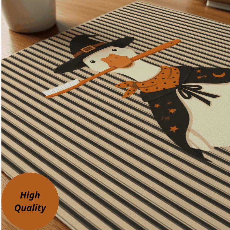 May include: A rectangular mat featuring a striped pattern in black and beige. The central graphic depicts a white goose wearing a black witch hat, an orange polka-dot bandana, and a black cape adorned with orange stars and a crescent moon.