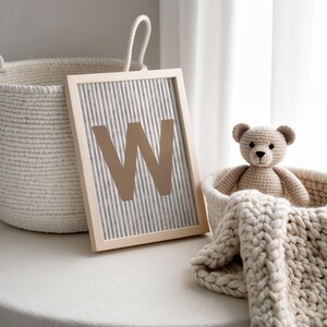 May include: A framed print with the letter "W" in a light brown color, set against a gray and white striped background. A woven basket and a crocheted teddy bear in a basket with a cream-colored blanket are also in the image.