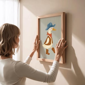 May include: A framed artwork featuring a cartoon duck dressed as a cowboy, wearing a blue hat, red bandana, and brown boots. The artwork has a light wood frame and a striped background. The image is being hung on a wall.
