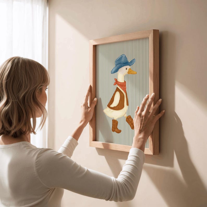 May include: A framed artwork featuring a cartoon duck dressed as a cowboy, wearing a blue hat, red bandana, and brown boots. The artwork has a light wood frame and a striped background. The image is being hung on a wall.