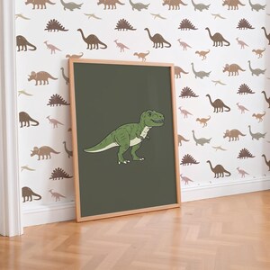 May include: A framed green Tyrannosaurus Rex illustration on a dark green background. The artwork is in a light wooden frame and leans against a wall with a dinosaur pattern. The floor is light brown.