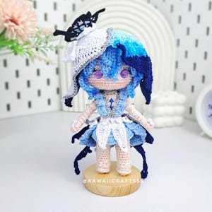 Può includere: Bambola all'uncinetto fatta a mano con capelli blu e un vestito abbinato. La bambola ha gli occhi viola e indossa un cappello a farfalla bianco e nero. La bambola è su una piccola base di legno. Il testo "@KAWAII CRAFTSSS" è sulla base.