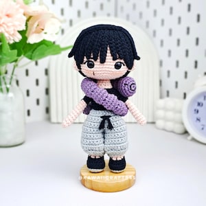 May include: A handcrafted crochet doll with black hair, a black top, grey shorts, and black shoes. A purple scarf is wrapped around the neck. The doll is displayed on a wooden base. The text "KAWAII CRAFTSSS" is visible on the base.