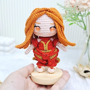Puede incluir: Muñeca de ganchillo hecha a mano con cabello naranja y un vestido rojo con detalles dorados. La muñeca tiene una cara rosa claro y está sobre una pequeña base de madera. Los ojos de la muñeca son naranjas. El texto "KAWAII CRAFTSSS" es visible.