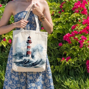 May include: A white canvas tote bag featuring a watercolour painting of a red and white lighthouse in the sea. The bag has a blue and white floral design.