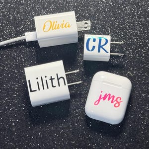 May include: Three white power adapters and one white wireless earbud charging case. The adapters feature the names Olivia in yellow, CR in blue, and Lilith in black. The charging case features the lowercase letters jms in pink.