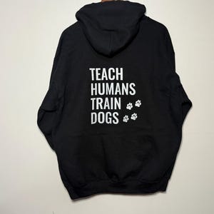 May include: Black hoodie featuring the white text "TEACH HUMANS TRAIN DOGS" and white paw prints. The sweatshirt has a hood and long sleeves. The background is a plain, off-white wall.