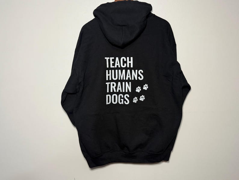 May include: Black hoodie featuring the white text "TEACH HUMANS TRAIN DOGS" and white paw prints. The sweatshirt has a hood and long sleeves. The background is a plain, off-white wall.