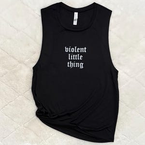 May include: Black muscle tank top with the phrase "violent little thing" in white gothic font. The sleeveless shirt is made of a soft material and is laying on a textured, off-white surface.