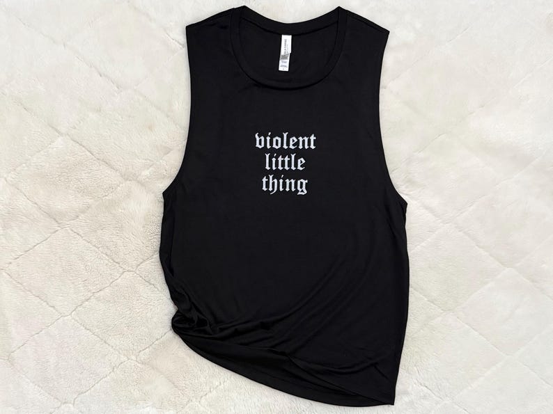 May include: Black muscle tank top with the phrase "violent little thing" in white gothic font. The sleeveless shirt is made of a soft material and is laying on a textured, off-white surface.