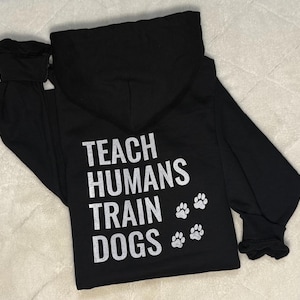 May include: Black hoodie featuring the text "TEACH HUMANS TRAIN DOGS" in white, accompanied by three paw prints. The hoodie includes a hood and long sleeves.