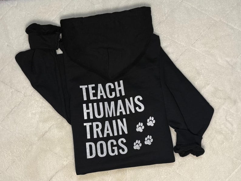 May include: Black hoodie featuring the text "TEACH HUMANS TRAIN DOGS" in white, accompanied by three paw prints. The hoodie includes a hood and long sleeves.