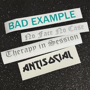 May include: Four rectangular adhesive vinyl decals with text. The top decal features teal text reading BAD EXAMPLE. Below are two decals with silver gothic-style text reading No Face No Case and Therapy in Session. The bottom decal has black text reading ANTISOCIAL.
