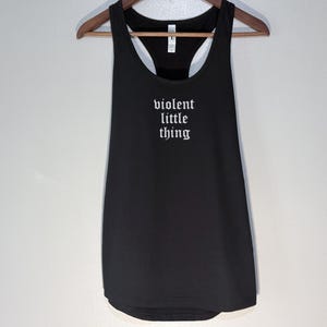 May include: Black tank top with the text "violent little thing" in white gothic font. The tank top is hanging on a wooden hanger against a white wall.