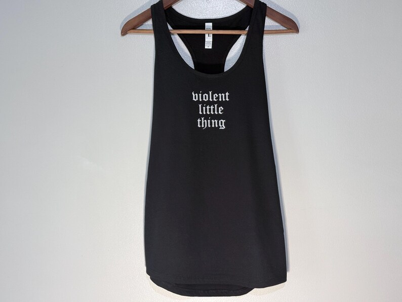 May include: Black tank top with the text "violent little thing" in white gothic font. The tank top is hanging on a wooden hanger against a white wall.