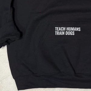 May include: Black hooded sweatshirt with the text "TEACH HUMANS TRAIN DOGS" in white block letters. The sweatshirt has a front pocket and ribbed cuffs. The fabric appears to be a soft, medium-weight material.