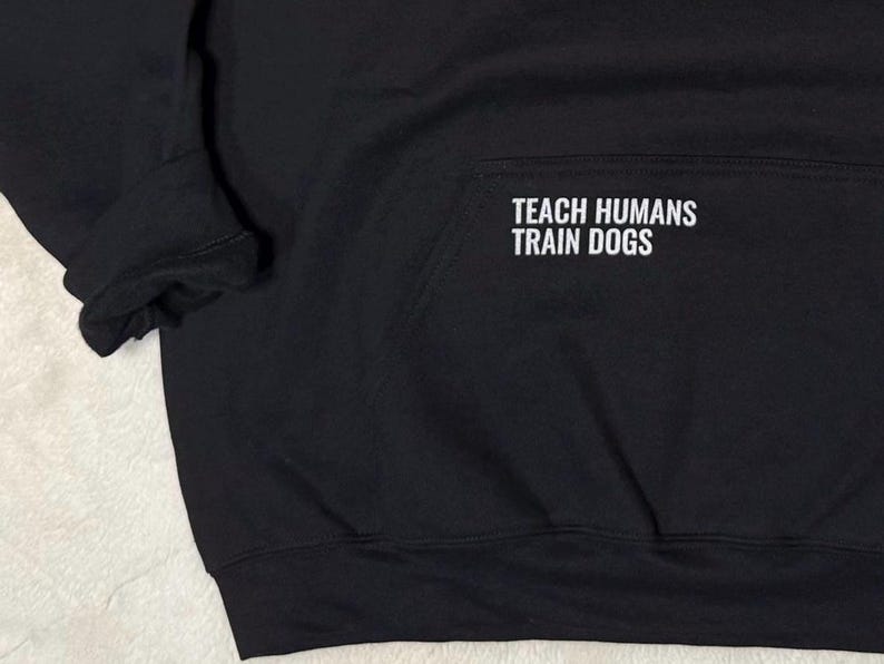 May include: Black hooded sweatshirt with the text "TEACH HUMANS TRAIN DOGS" in white block letters. The sweatshirt has a front pocket and ribbed cuffs. The fabric appears to be a soft, medium-weight material.