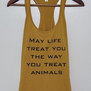 May include: A mustard yellow racerback tank top featuring the black text "MAY LIFE TREAT YOU THE WAY YOU TREAT ANIMALS" printed on the front. The garment has a scoop neckline and is displayed on a wooden hanger against a plain white background.