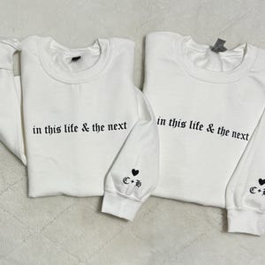 May include: Two white sweatshirts with the phrase "in this life & the next" printed in black. Each sleeve has a heart and the initials "C+B". The sweatshirts are a matching set.