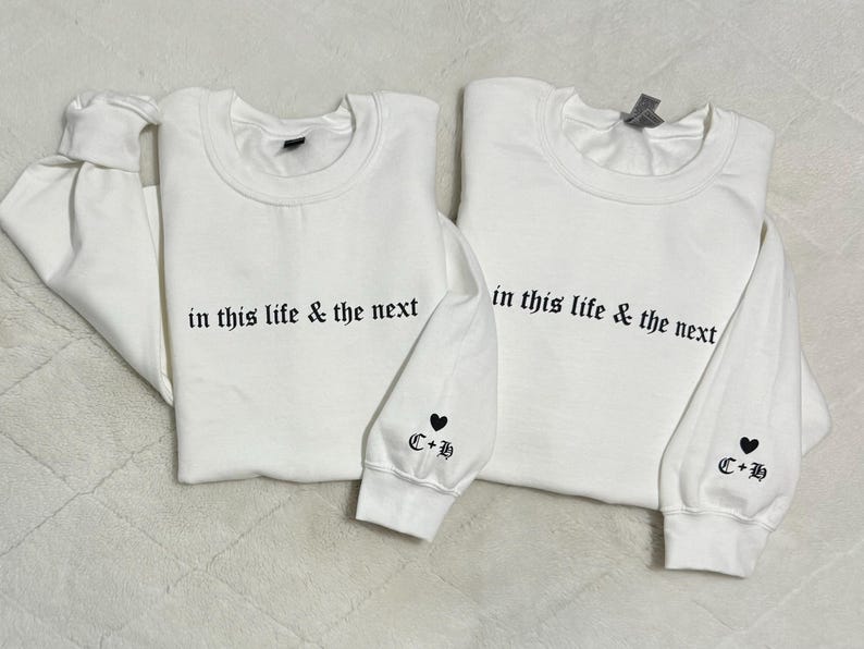 May include: Two white sweatshirts with the phrase "in this life & the next" printed in black. Each sleeve has a heart and the initials "C+B". The sweatshirts are a matching set.