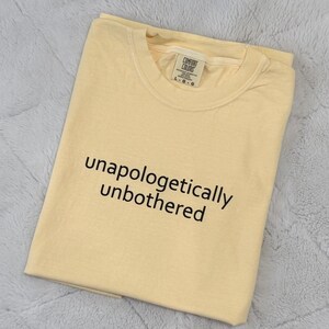 May include: A light yellow crew neck t-shirt featuring the black text "unapologetically unbothered" printed on the front. The shirt has a Comfort Colors brand label visible at the inner neckline, indicating a size L, and short sleeves.