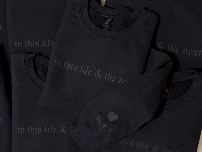 May include: Black crewneck sweatshirts with the phrase "in this life & the next" printed in a gothic font. One sweatshirt sleeve has a heart graphic and the initials "B+C".