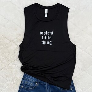 May include: Black sleeveless tank top with the text "violent little thing" in a gothic font. The tank top is paired with blue denim jeans. The image is taken on a white, textured surface.