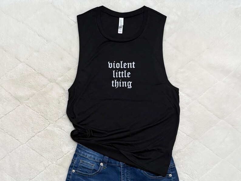 May include: Black sleeveless tank top with the text "violent little thing" in a gothic font. The tank top is paired with blue denim jeans. The image is taken on a white, textured surface.