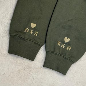 May include: Close-up of two olive green sweatshirt sleeves. Each sleeve cuff features gold embroidered initials and a heart. The initials are "N&A" on one sleeve and "A&N" on the other. The cuffs are ribbed.