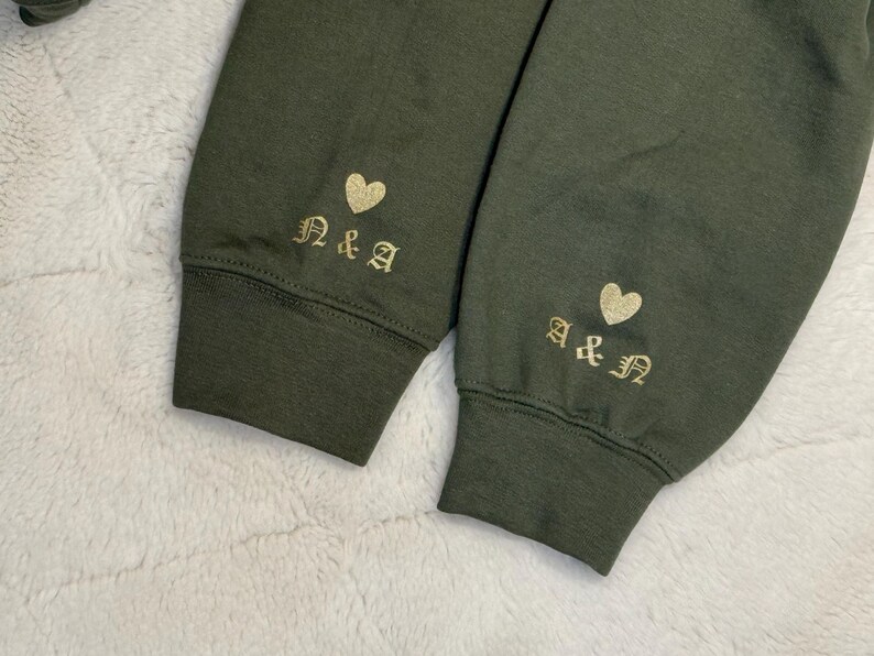 May include: Close-up of two olive green sweatshirt sleeves. Each sleeve cuff features gold embroidered initials and a heart. The initials are "N&A" on one sleeve and "A&N" on the other. The cuffs are ribbed.