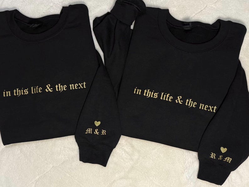 May include: Two black sweatshirts with gold embroidered text that reads "in this life & the next." One sleeve has "M&R" and a heart, the other has "R&M" and a heart. The sweatshirts are on a white surface.
