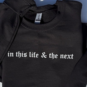 May include: Black sweatshirt with the phrase "in this life & the next" in white gothic font. A small white heart and initials are visible on the sleeve. The Gildan tag is visible on the collar.