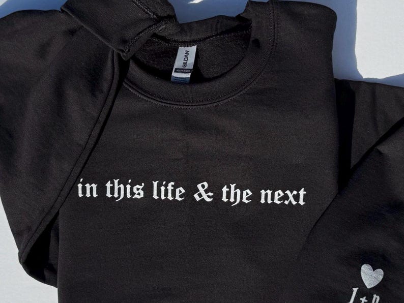 May include: Black sweatshirt with the phrase "in this life & the next" in white gothic font. A small white heart and initials are visible on the sleeve. The Gildan tag is visible on the collar.