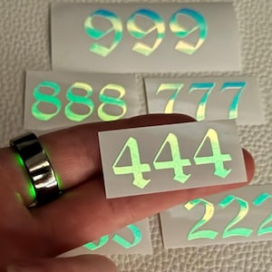 May include: A collection of iridescent number decals, including 999, 888, 777, 444, and 222. A black ring with a glowing green interior is also visible. The numbers have a holographic effect, shifting colors.